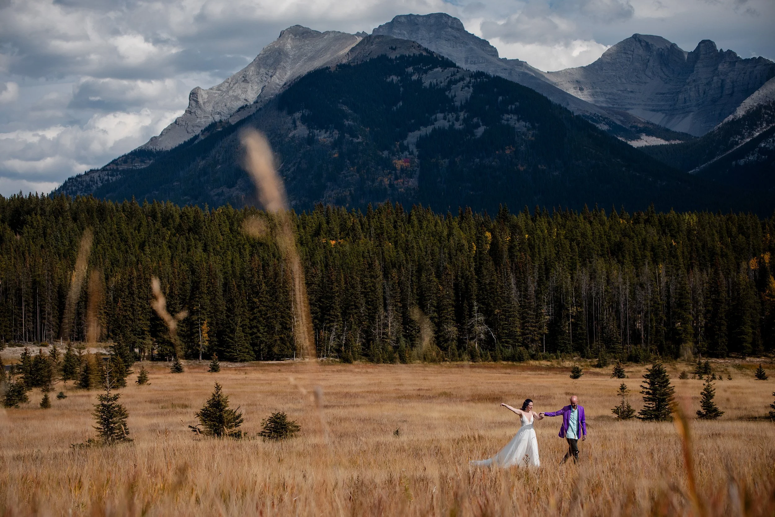 Banff Wedding in September. Couple Walks in field with large mountains and heavy clouds behind them. 