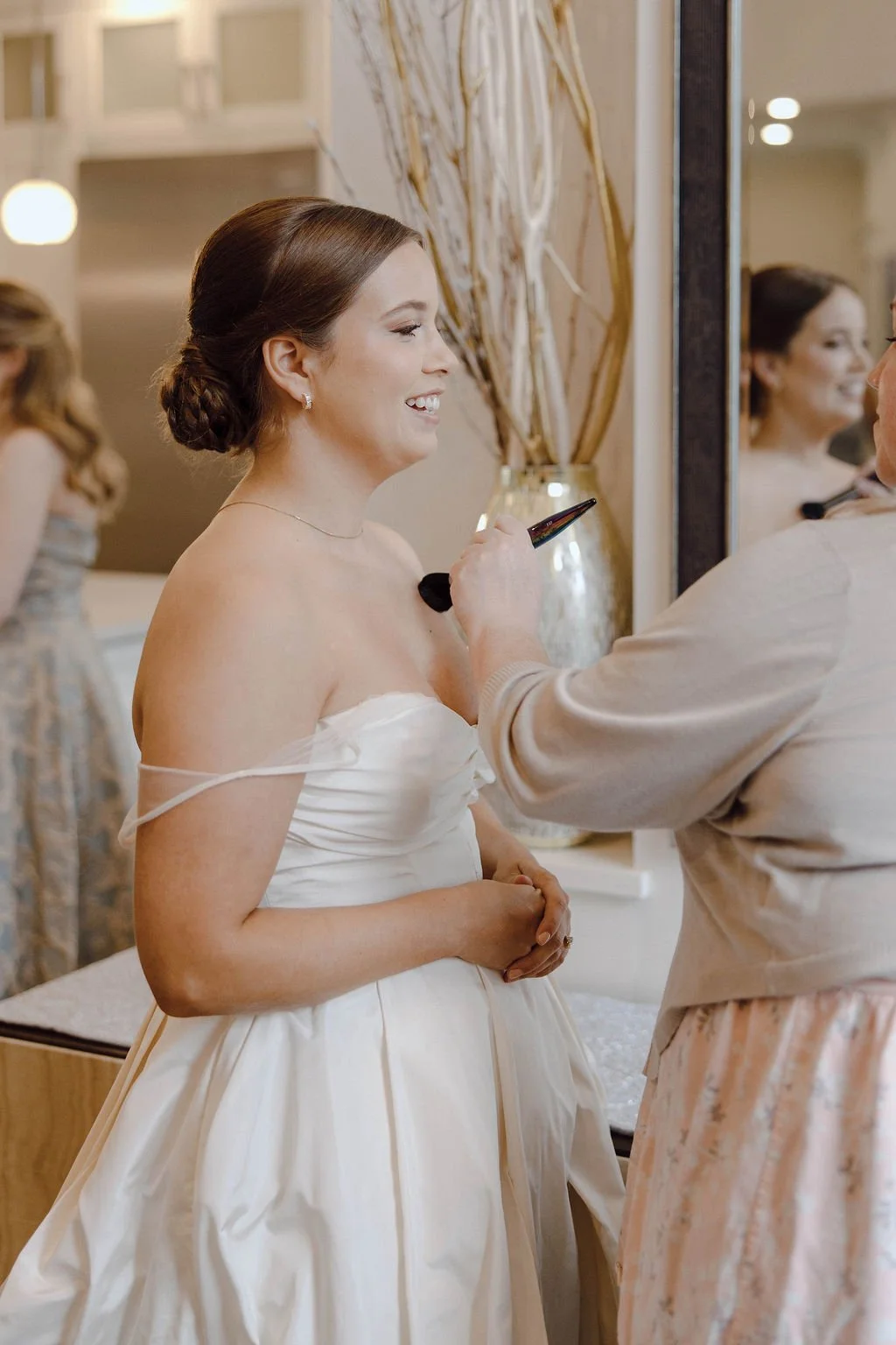 A woman in an off-shoulder white dress having makeup applied by a makeup artist in a well-lit room decorated with a large vase of dried decorative branches and a mirror.