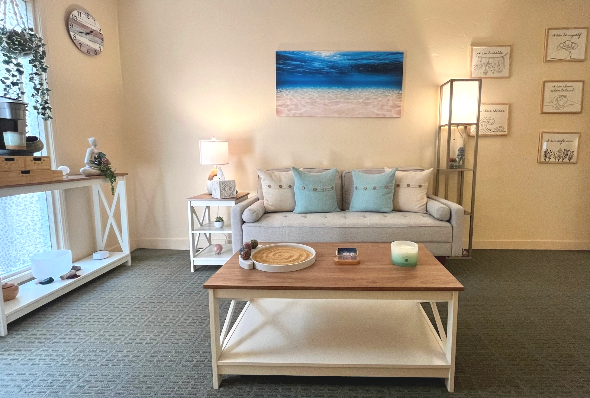 Living room with a white sofa, blue pillows, wooden coffee table with a tray and candle, wall art depicting a beach, and decorative items including sculptures and framed prints.