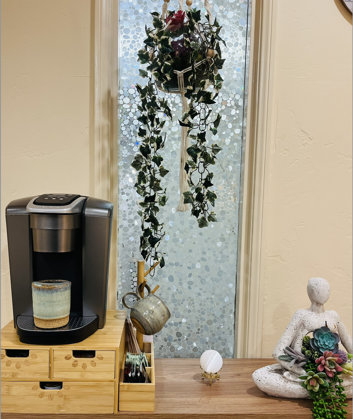 A coffee station with a Keurig machine, a ceramic mug, and a wooden organizer with drawers and compartments. Behind it is a decorative frosted window with a hanging ivy plant in a wall-mounted planter and a small white orb light on the desk. To the r