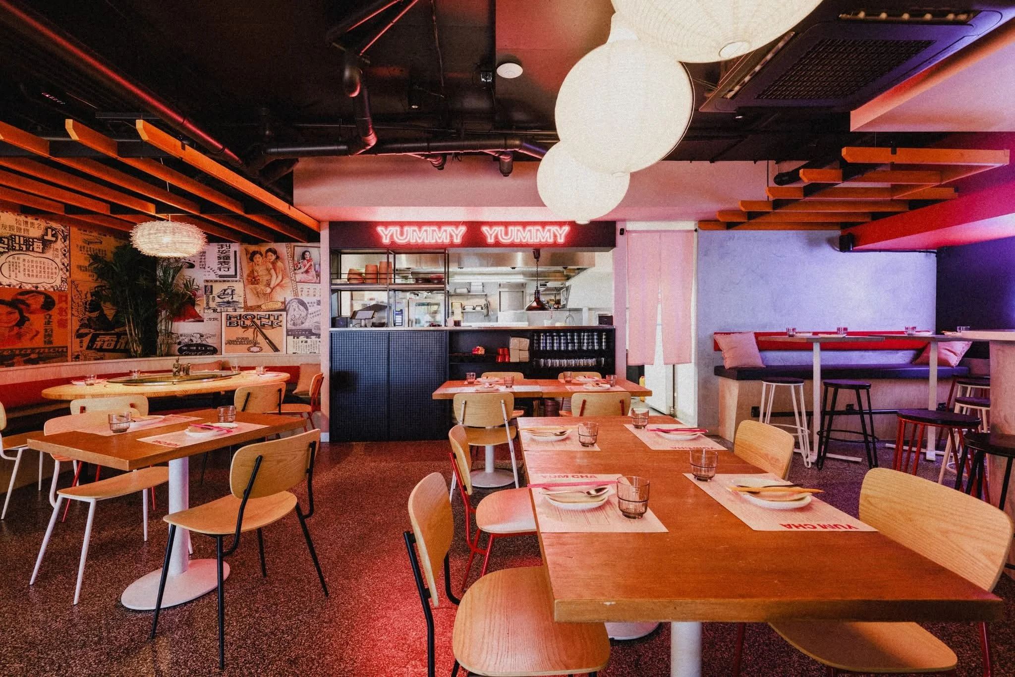 Interior of a modern restaurant with wooden tables, chairs, and a bar area. Decor includes vintage posters on the wall, large white spherical hanging light fixtures, and a neon sign that says 'YUMMY YUMMY'.