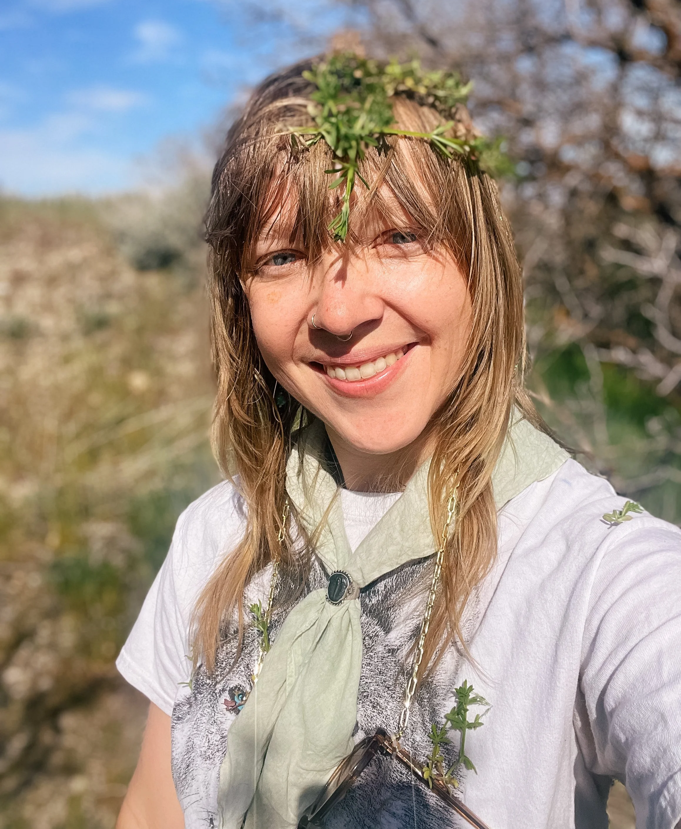 A smiling woman with a small green plant on her forehead outdoors on a sunny day with trees and blue sky in the background.
