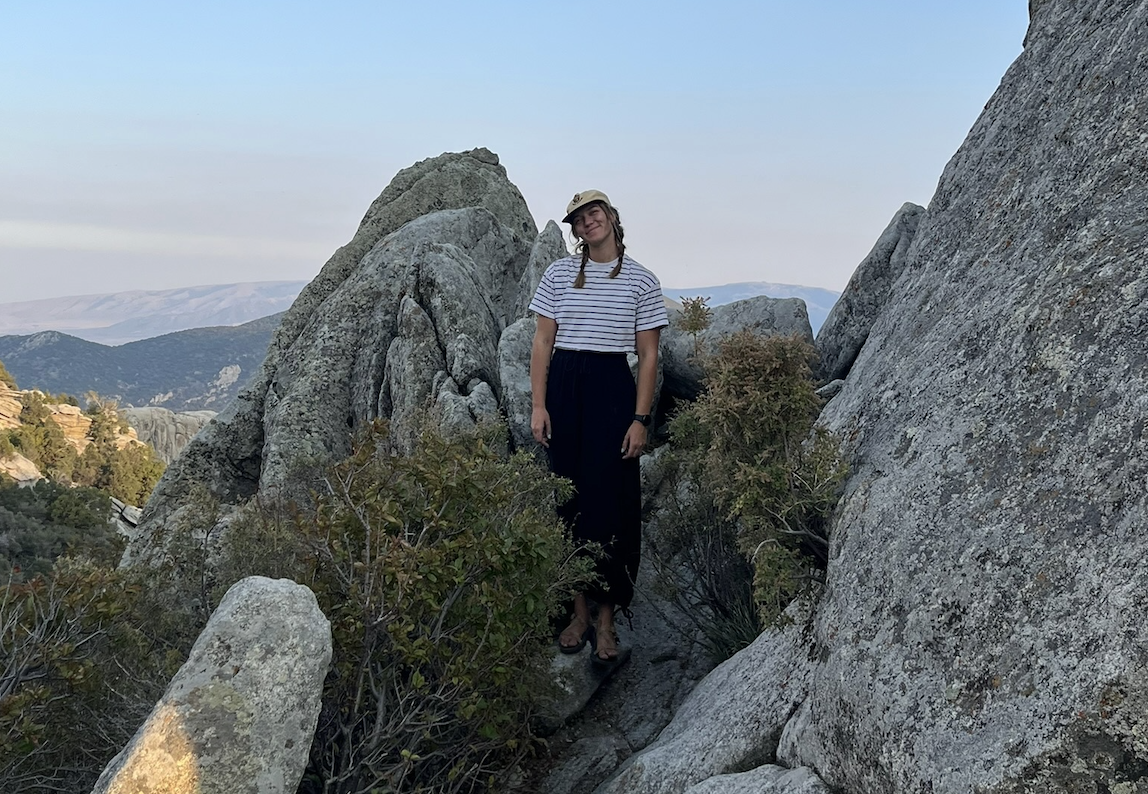 Woman in striped shirt and wide-brim hat standing among large rocks and bushes in a mountainous outdoor setting.