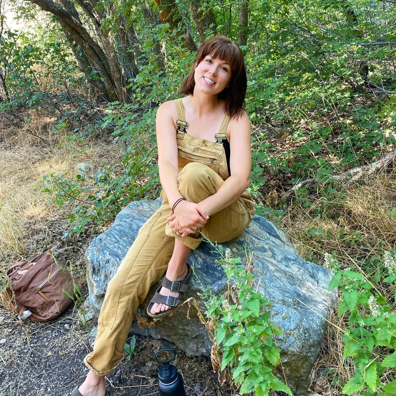 A woman sitting on a large rock in a forest, smiling with short brown hair, wearing tan overalls, black tank top, and sandals, with a brown backpack nearby.