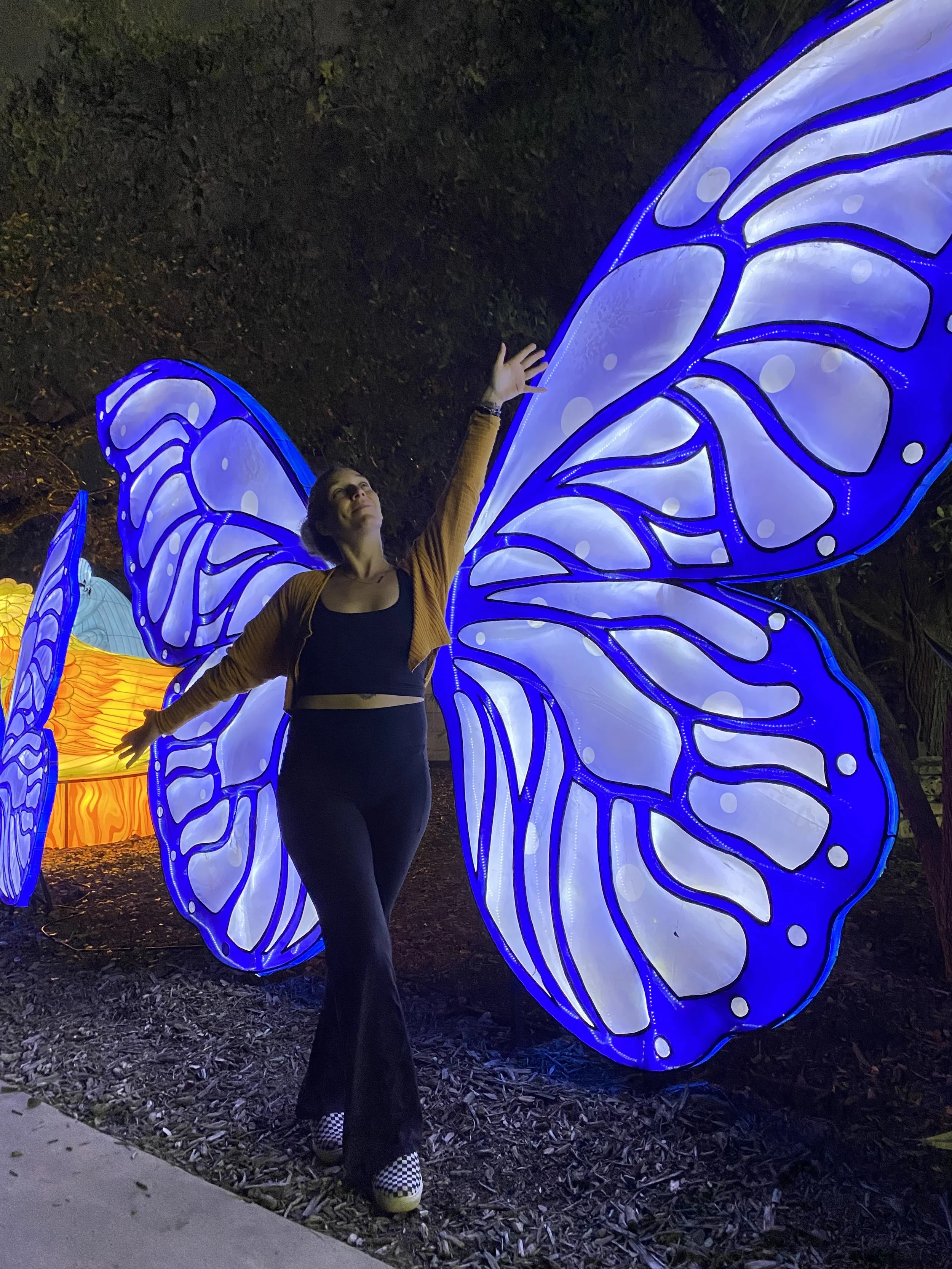 A woman standing with arms raised in front of illuminated blue butterfly-shaped lanterns at night.