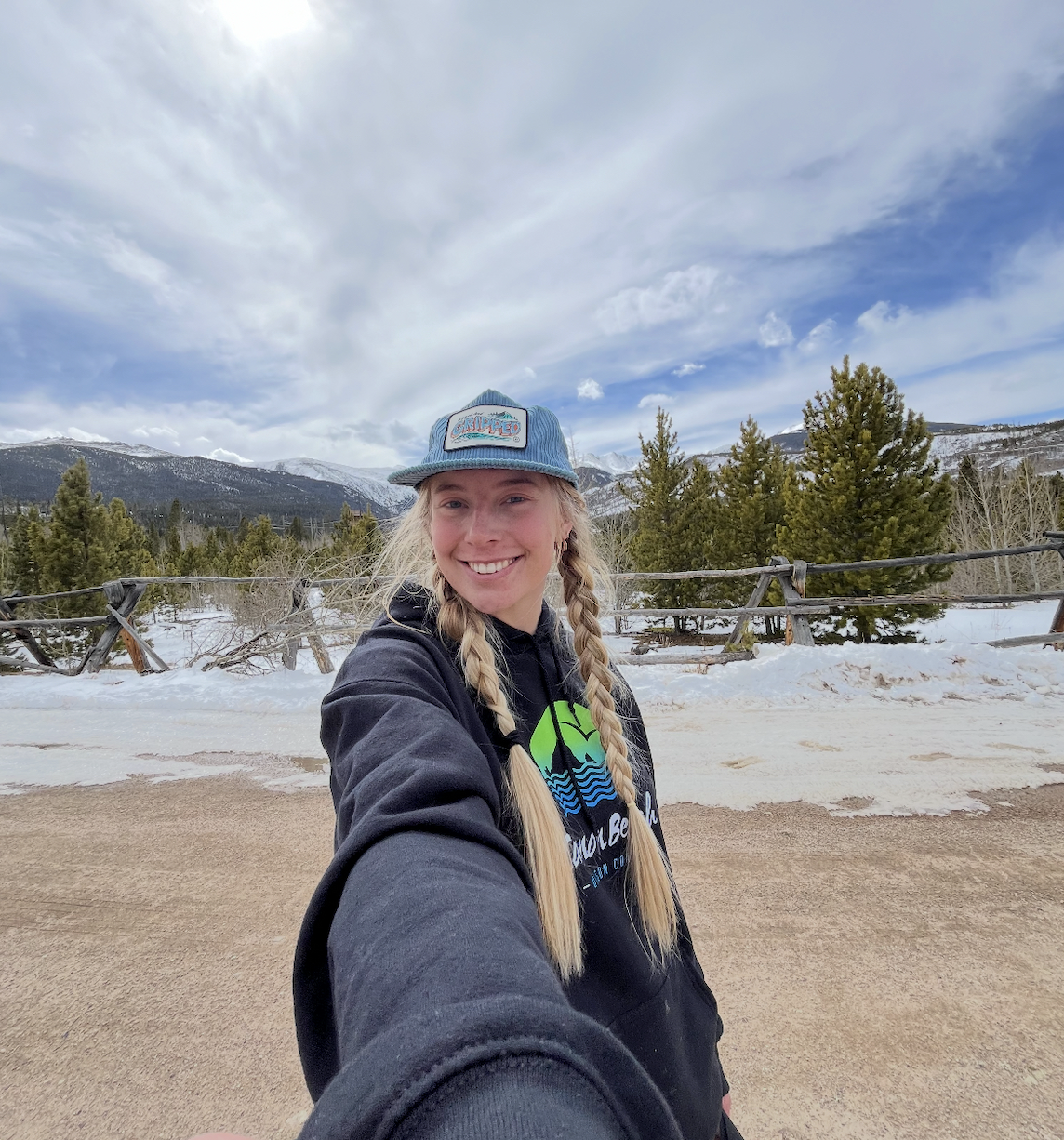 A young woman with blonde braids wearing a blue cap and black hoodie, taking a selfie in a snowy mountainous landscape with trees and a cloudy sky.