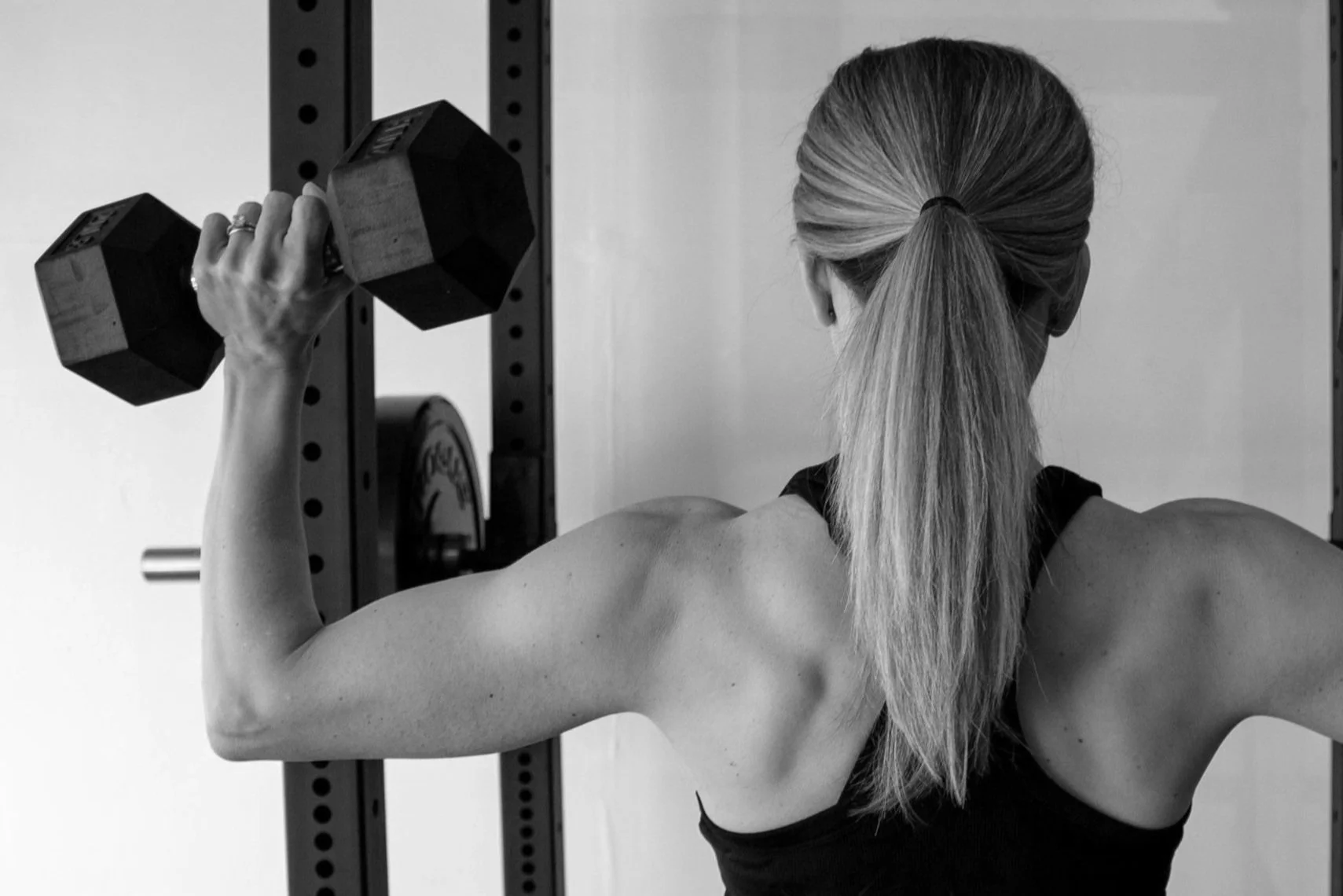 A woman with blonde hair smiling, crossing her arms, standing in a gym near weightlifting equipment.