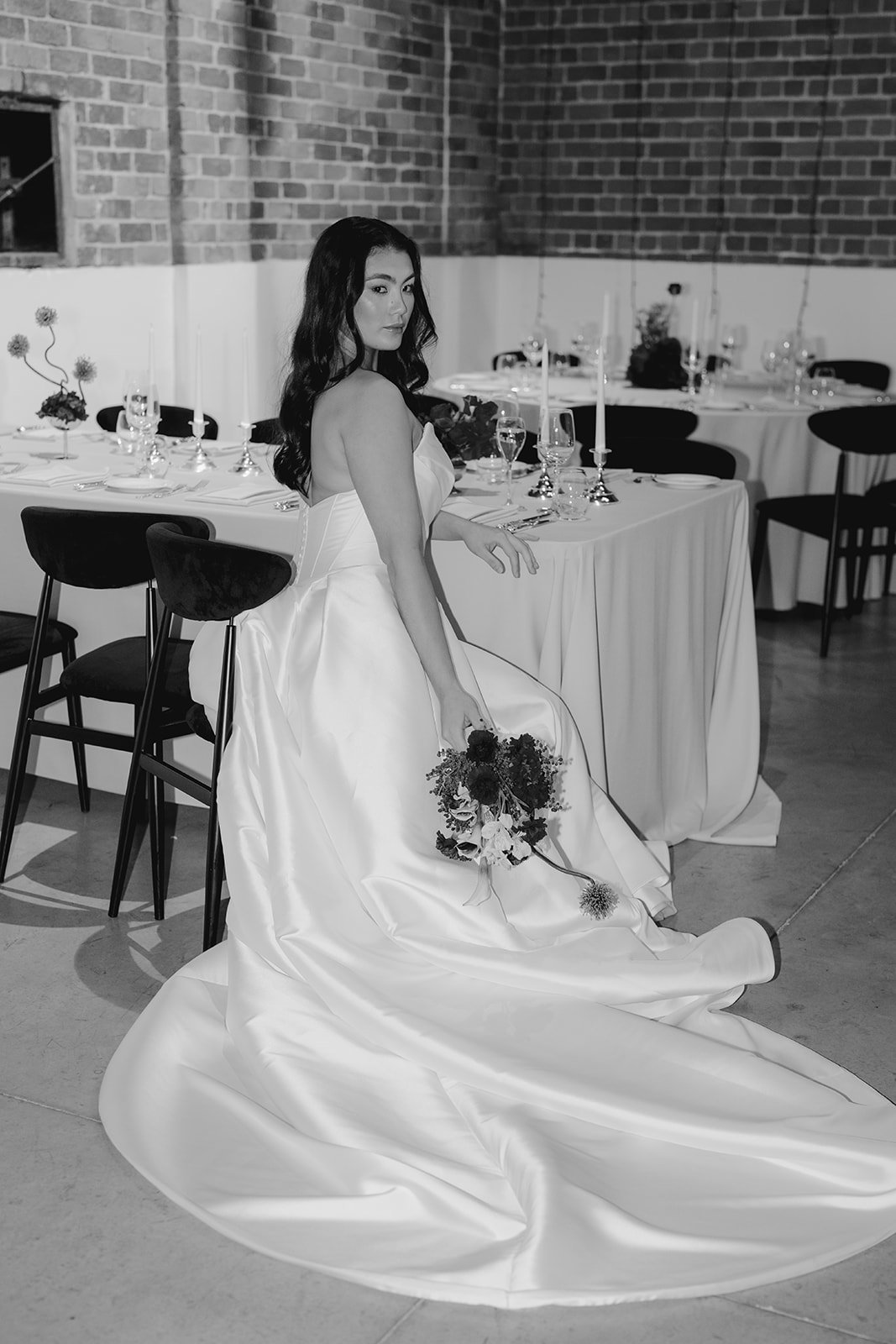 Black and white photo of a bride seated at a rectangular banquet table at Nikola Estate wedding reception, highlighting elegant table styling, structural florals, and the luxurious, moody atmosphere of the rustic Old Cellars venue.
