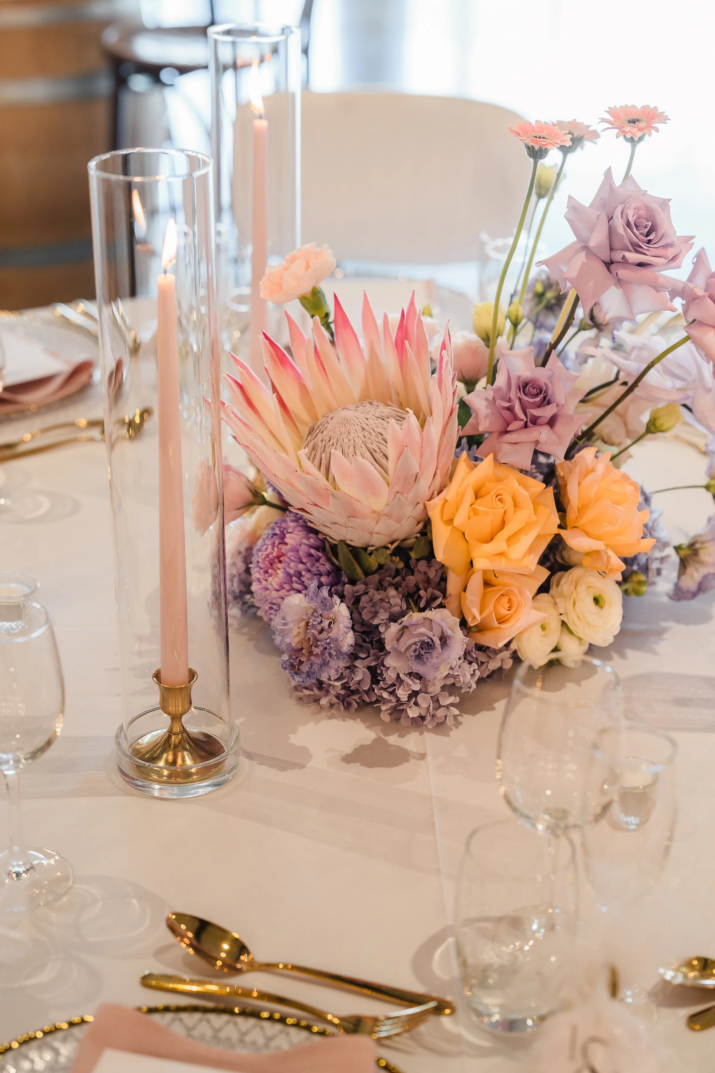 Soft, whimsical floral arrangements with pink king protea, blush taper candle in gold holder and glass sleeve, gold-accented table styling with blush napkins