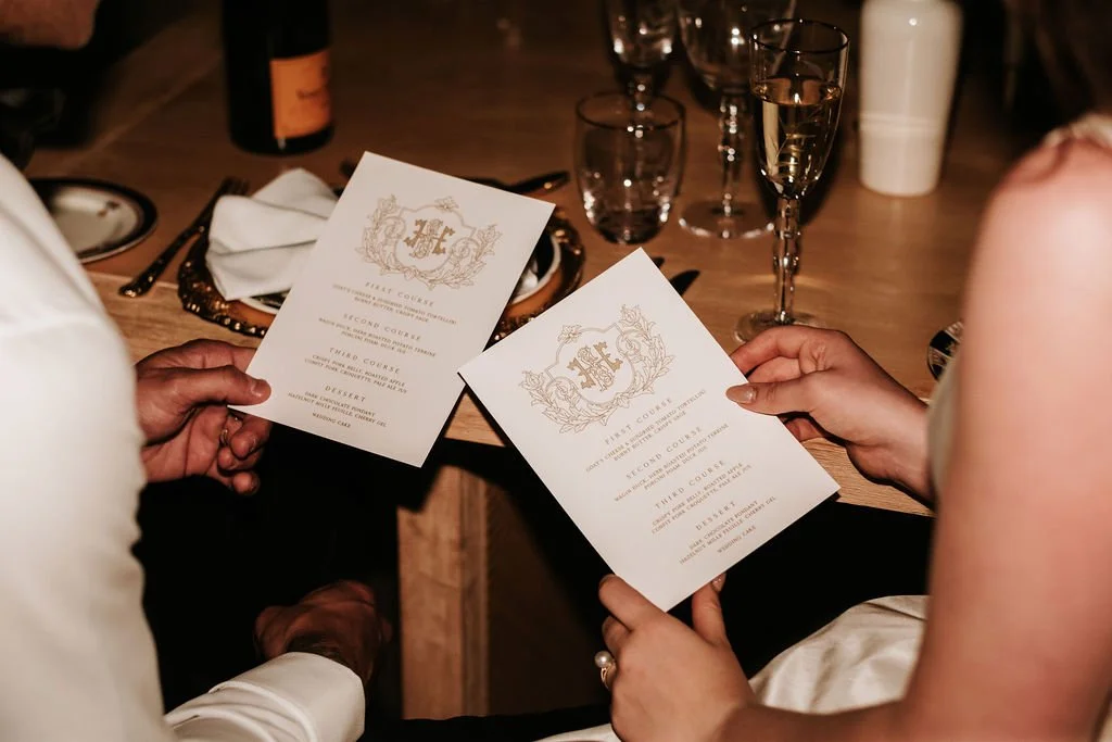 Over shoulder shot of bride (right) and groom (left) seated at wedding reception table holding custom designed white and gold menu by Paper and Ink Studios, bottle of Veuve Clicquot atop wooden table in background, romantic place setting, luxury plac