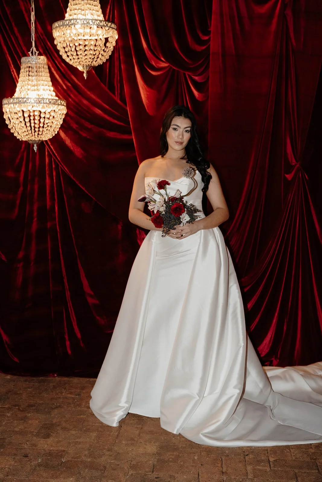 Ceremony setup in Nikola Estate’s Moondyne Joe Room featuring paved red brick flooring, rich red velvet draping behind the bride in a voluminous white wedding dress and hanging crystal chandeliers, with deep purple, white and red florals bouquet.