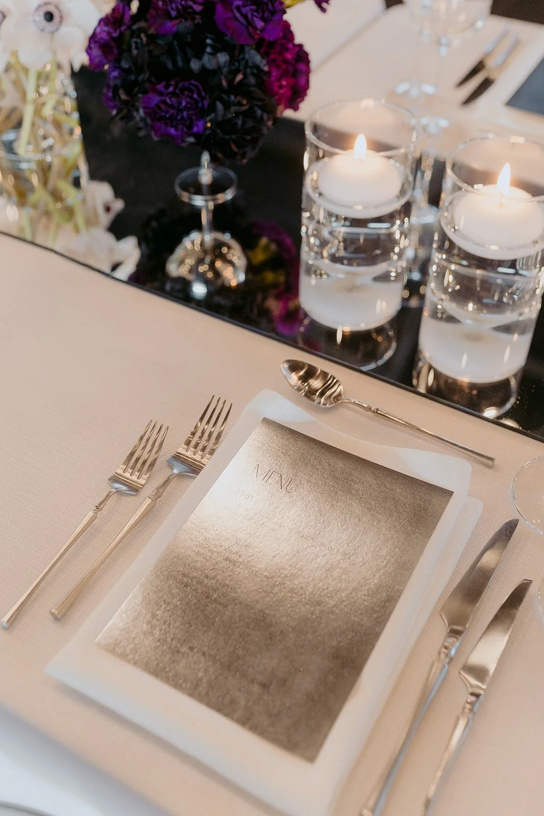 Sophisticated wedding guest place setting featuring white linens, statement silver cutlery, floating candles and deep purple and white florals set atop a mirrored runner.