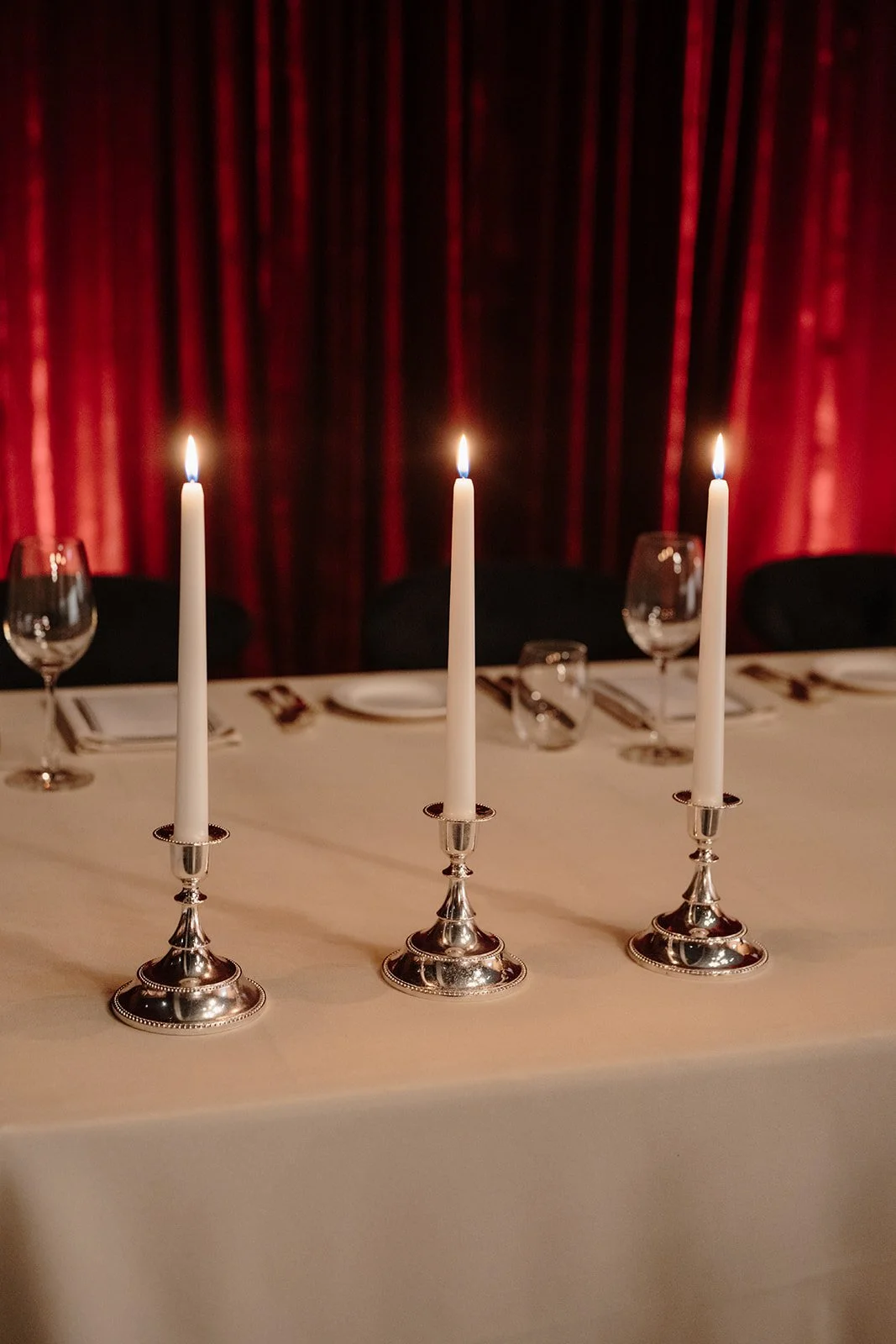 Buring white candles in vintage silver holders on a floorlength tan tablecloth, with refined place-settings in the background as well as rich red velvet draping and uplighting at Nikola Estate, Swan Valley wedding reception.