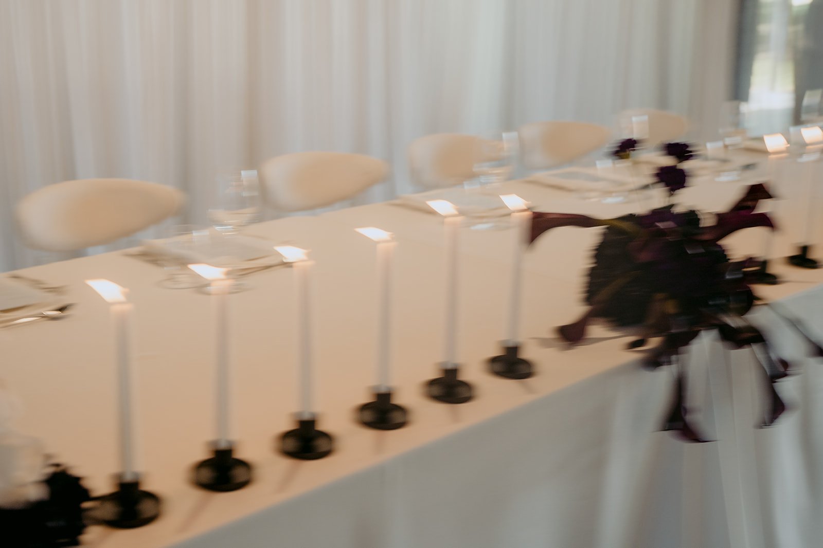 Bridal table with textured white linens, taper candles, deep purple florals and a draped backdrop.