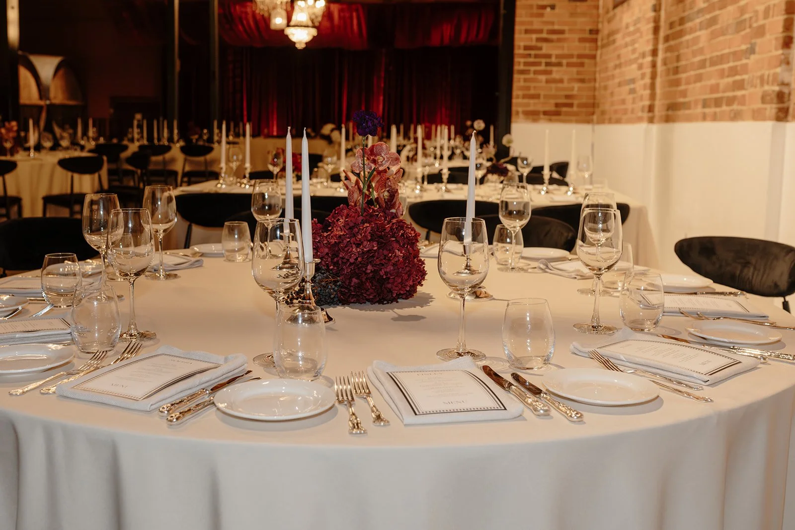 Bold and luxurious wedding reception at Nikola Estate’s Old Cellars in the Swan Valley, featuring dramatic red draping, black velvet chairs, white and silver accents and a tan, deep purple, and red colour palette for an elegant 80-guest celebration.