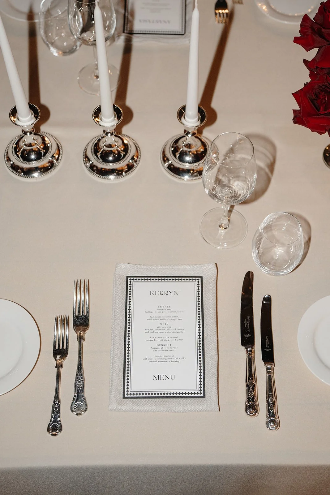 High-end, elegant wedding place setting at Nikola Estate, Swan Valley with black bordered white menu, vintage silver cutlery and candle holders, white side plate, modern stemware and structural red rose floral arrangement, with tan-on-tan napkin and 