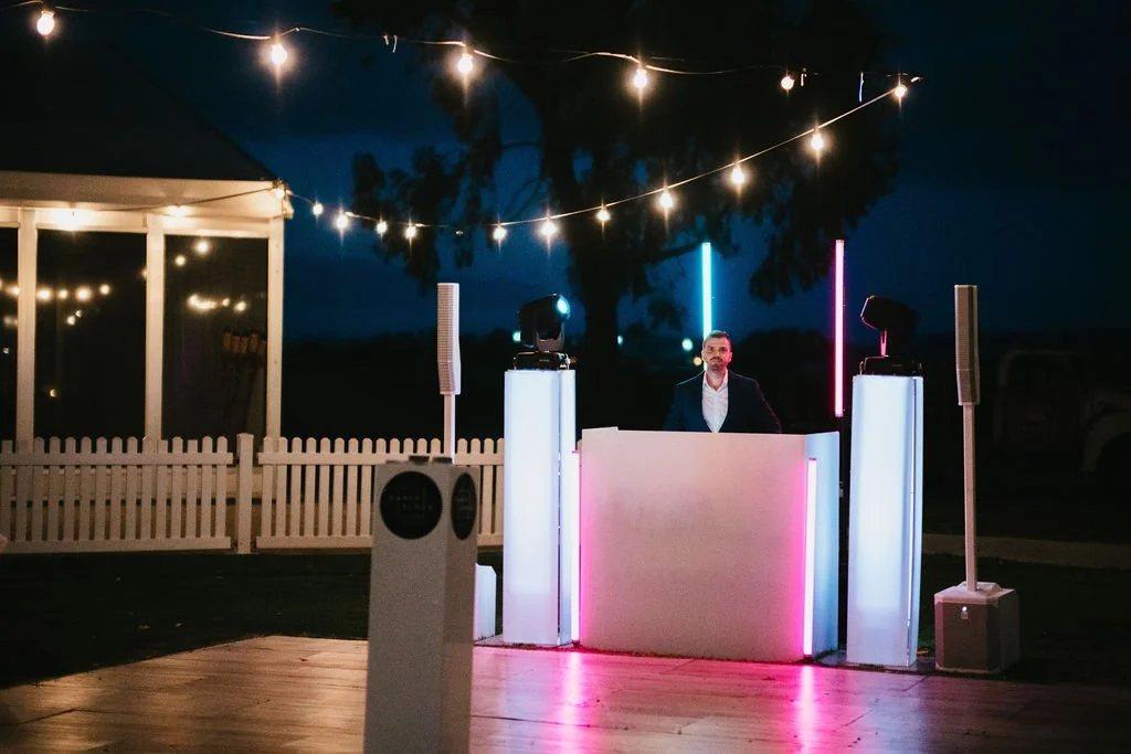 DJ booth at night lit up with overhead festoon lights and uplights, white ash dancefloor and white acrylic DJ setup by Complete DJ, white marquee and picket fence in background to left of image, Swan Valley Wedding, modern wedding, white wedding