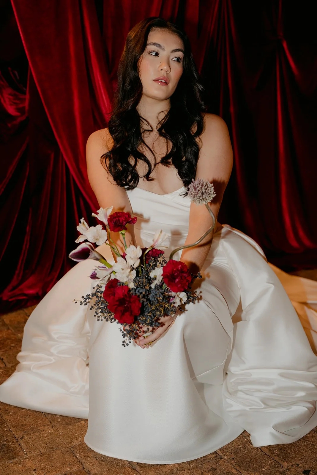 Ceremony setup in Nikola Estate’s Moondyne Joe Room featuring paved red brick flooring, rich red velvet draping behind the bride in a voluminous white wedding dress with deep purple, white and red florals bouquet.