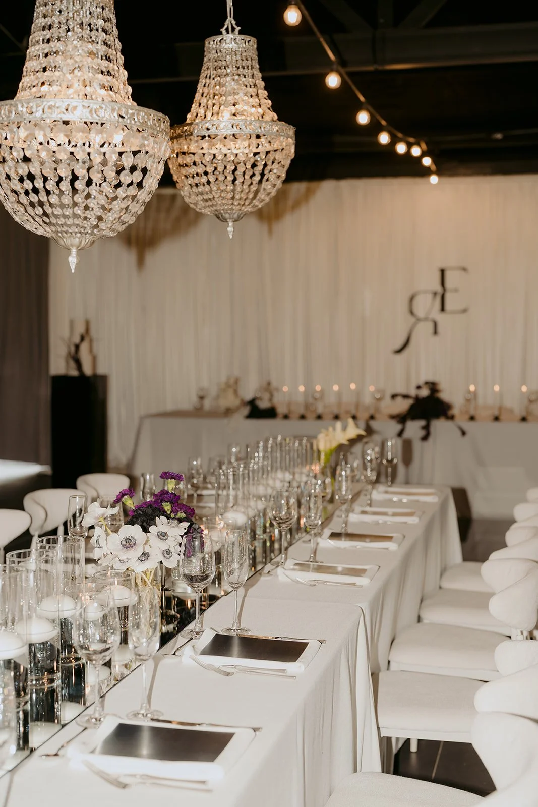 Contemporary wedding tablescape with textured white linens, mirror table runner, silver details, elegant place settings, floating candles and chandeliers set against a draped backdrop with black monogram.