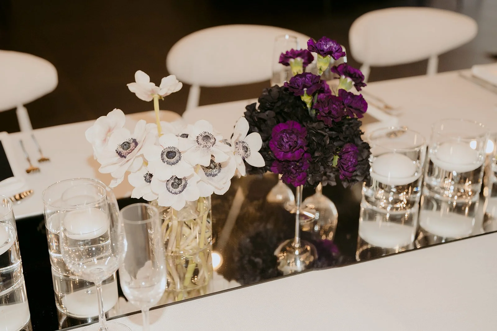 Contemporary wedding tablescape with textured white linens, floating candles and white and deep purple florals set atop a central mirrored runner reflecting the chandeliers overhead and sophisticated place settings with reflective menus and statement