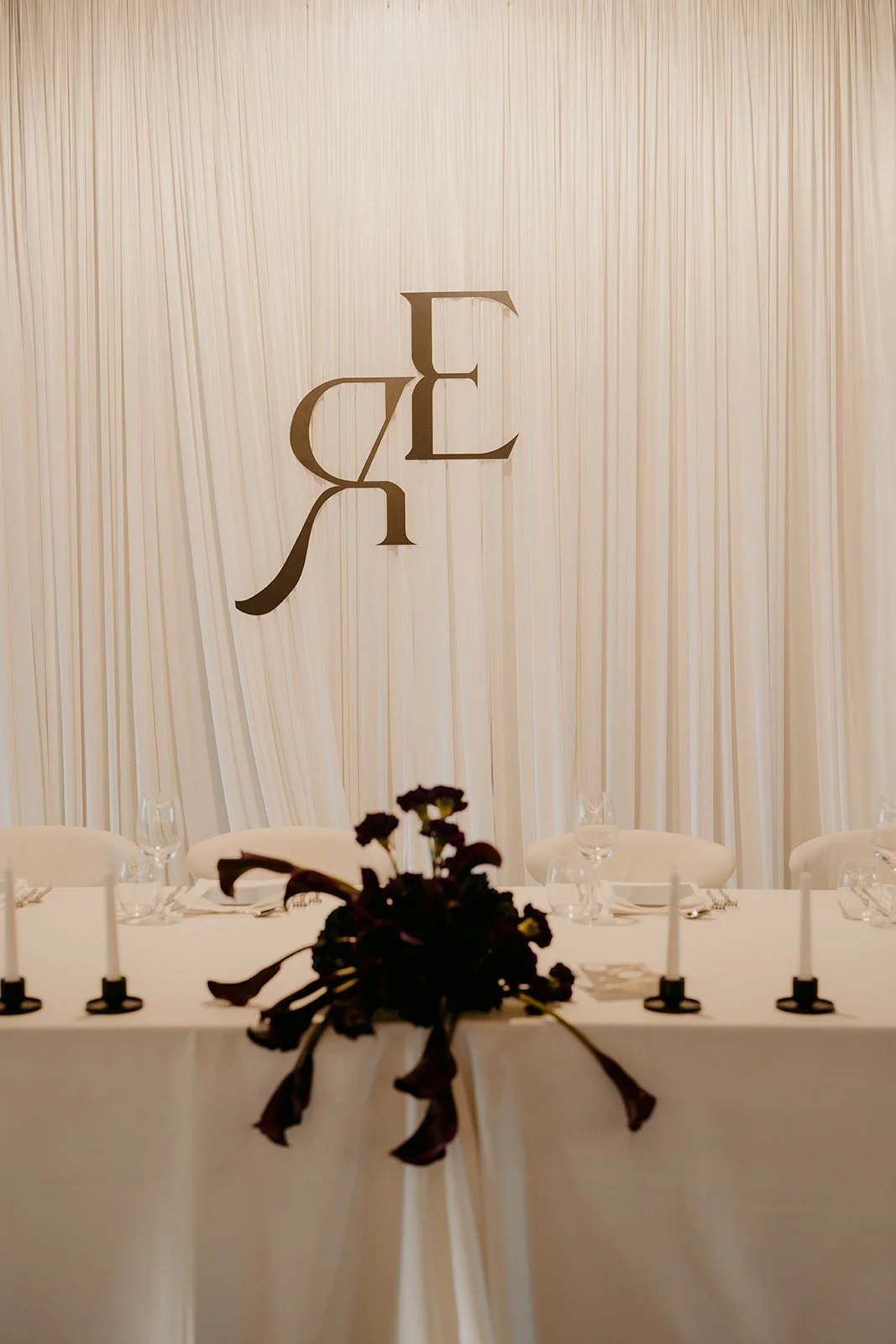 Bridal table featuring a draped backdrop, textured white linens, black monogram, deep purple florals and candles.