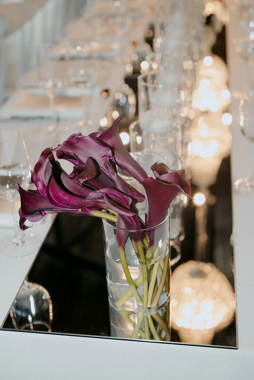 Contemporary wedding tablescape with textured white linens, floating candles and deep purple florals set atop a central mirrored runner reflecting the chandeliers overhead and sophisticated place settings with reflective menus and statement silver cu