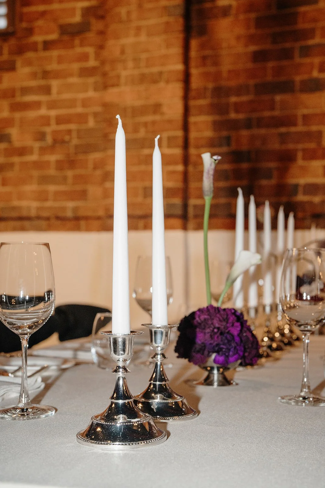 High-end wedding table styling at Nikola Estate with rectangular banquet table, elegant place settings with silver accents and structural deep purple and white floral arrangement, with tan-on-tan napkin and tablecloth.