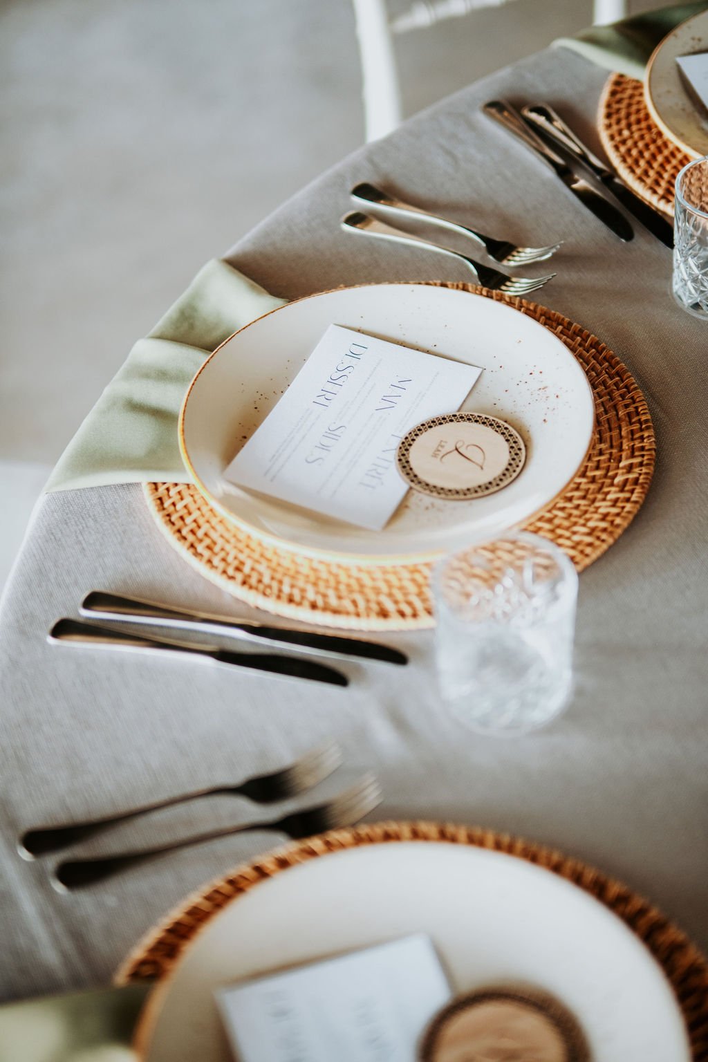 Image showing three place settings of round banquet table seating eight adorned with dove grey linen tablecloth, dusty green linen napkins draped over brown rattan placemats and over edge of table at each setting, brown speckled off-white plates sit
