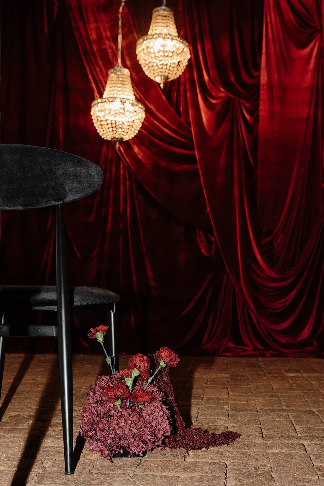 Ceremony setup in Nikola Estate’s Moondyne Joe Room featuring black velvet chairs, red brick walls, paved brick flooring, rich red velvet draping and hanging crystal chandeliers, with deep purple and red florals along the aisle.