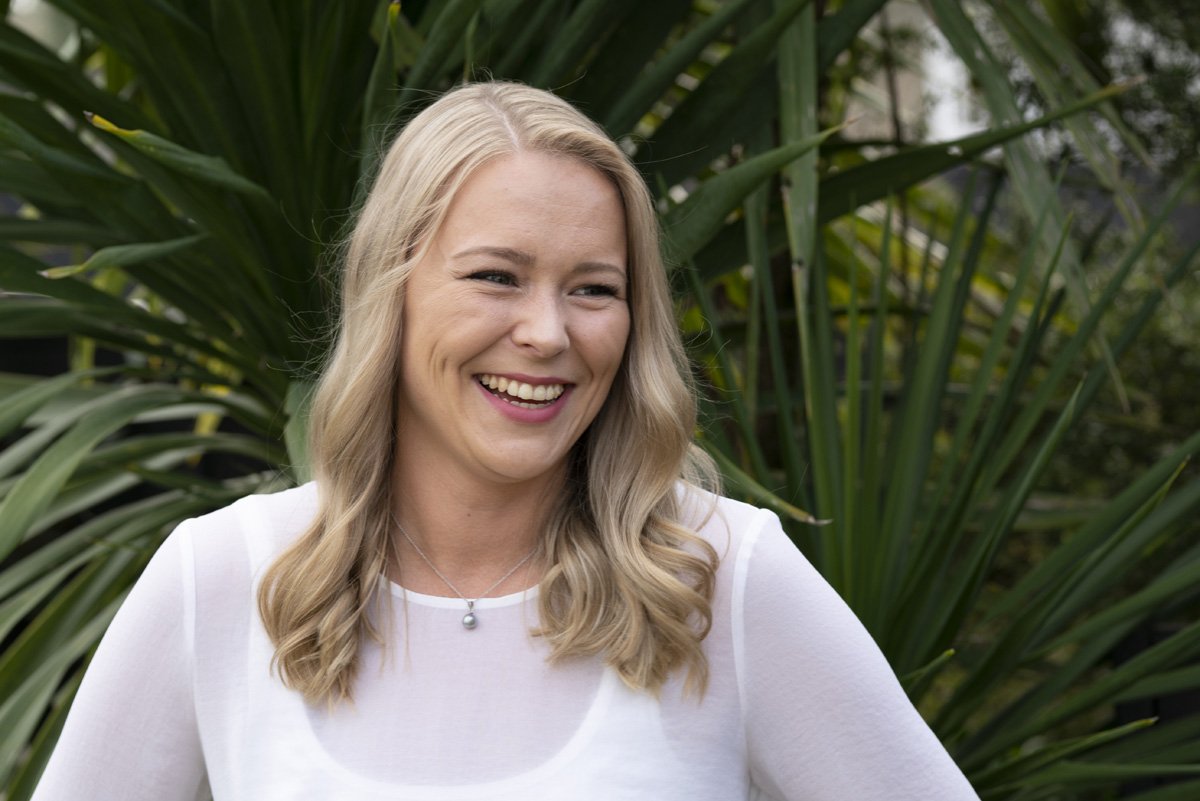 Smiling woman with long blonde hair wearing a white top, standing in front of green leafy plants.