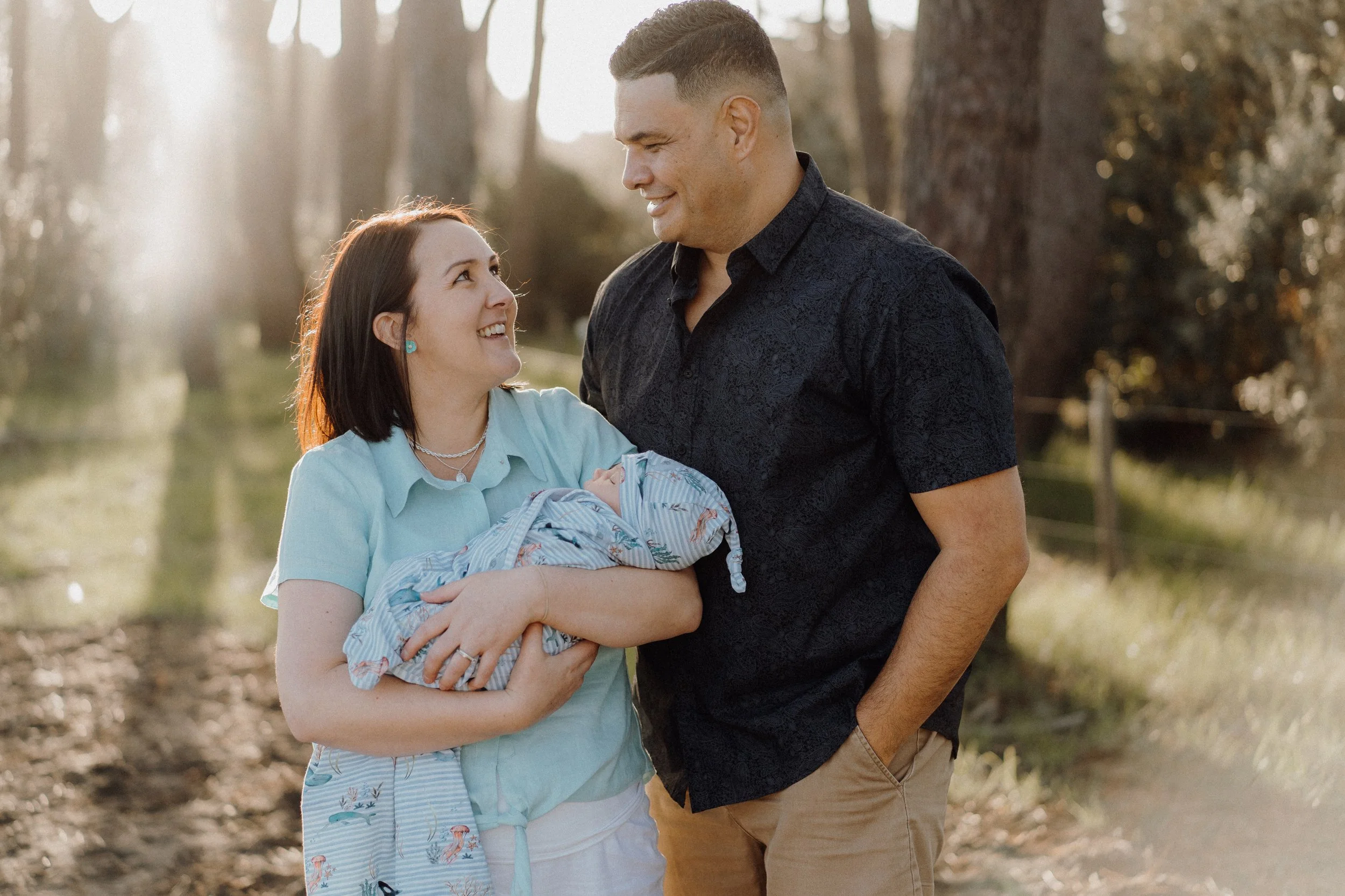 A woman holding a sleeping newborn baby outdoors, smiling at a man, with trees and sunlight in the background.