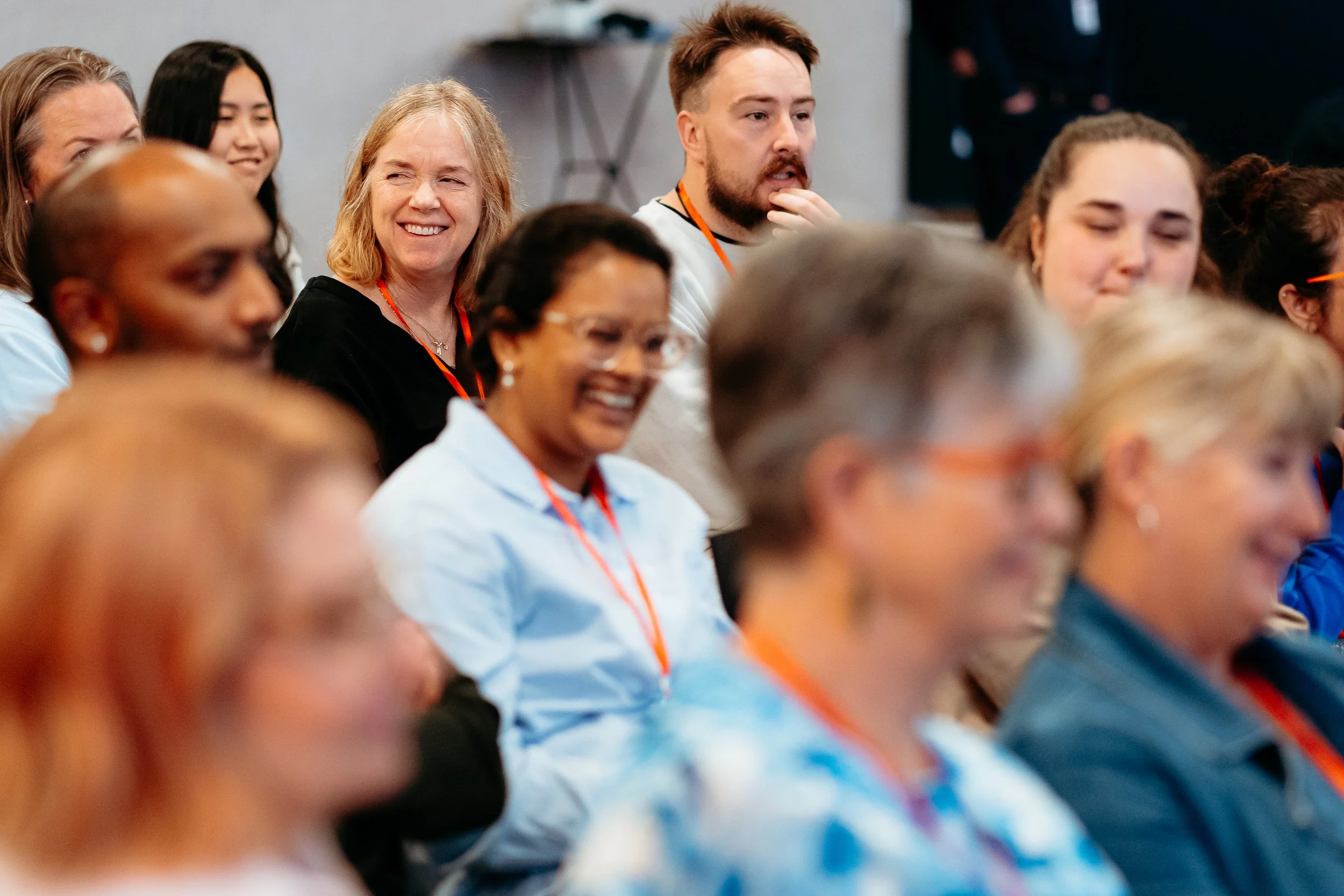 Group of diverse adults attending a conference, seated and listening attentively, some smiling and engaging with the speaker or presentation.