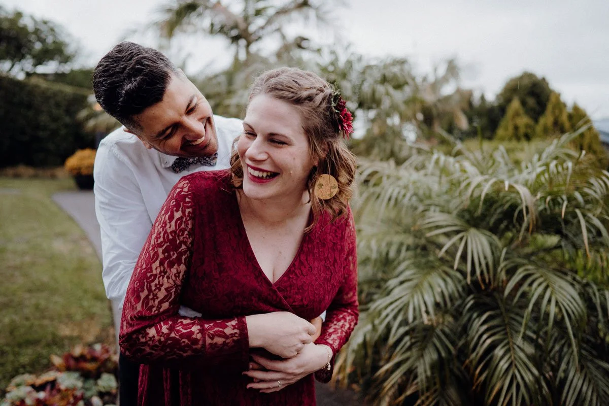A man and woman laughing outdoors, the man hugging the woman from behind, both smiling.