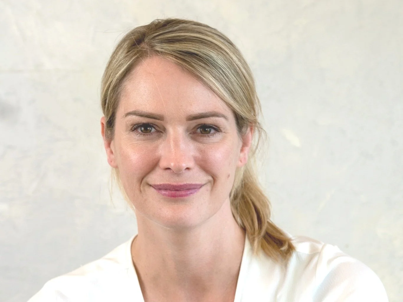 A woman with blonde hair in a white shirt, smiling slightly, sitting against a light-colored wall.
