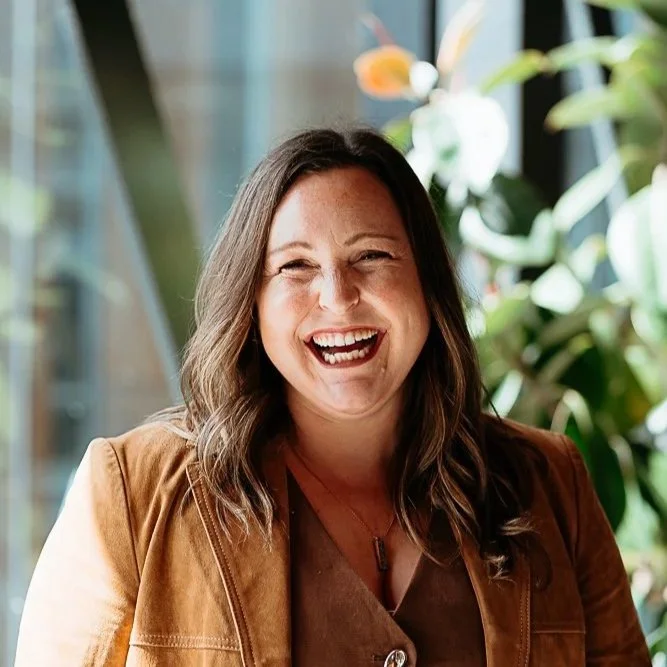 A woman with shoulder-length brown hair laughing in an indoor setting with green plants in the background.