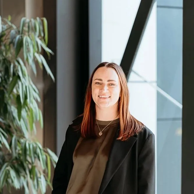 A woman with brown hair smiling indoors near large windows and a green leafy plant.