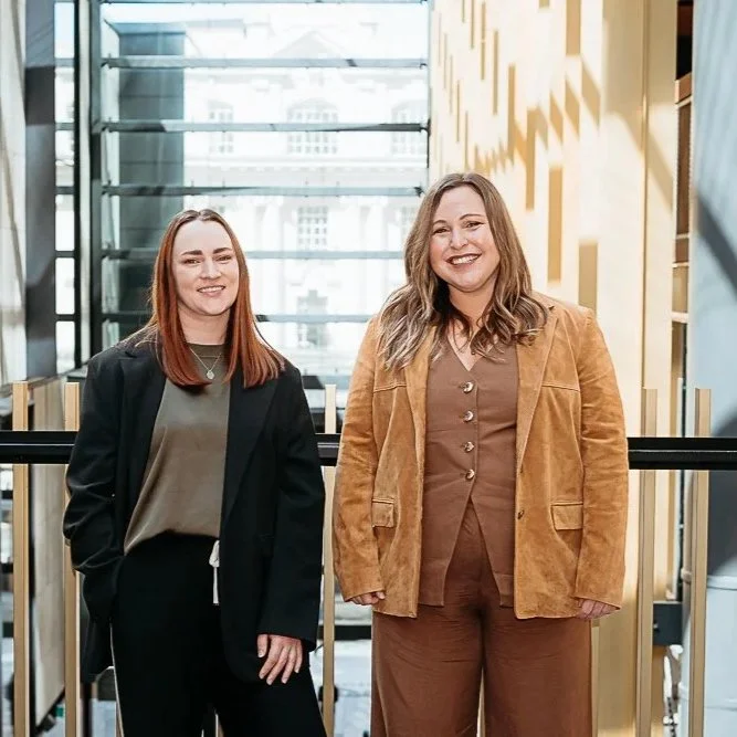 Two women standing side by side in an indoor modern building with glass walls. They are smiling and dressed in business casual attire.