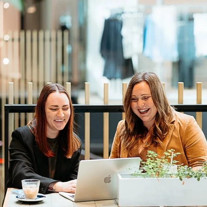 Two women sitting at a table, laughing and looking at a laptop, with a coffee cup and a potted plant on the table in a retail store or cafe.