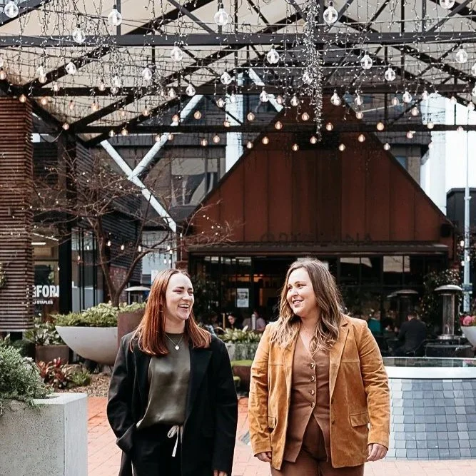 Two women smiling and talking outdoors under a string of hanging lights. They are standing in front of a building with a covered porch, and there are plants and trees around them.