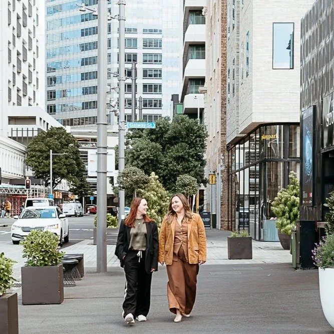 Two women walking and talking on a city sidewalk surrounded by modern buildings and greenery.