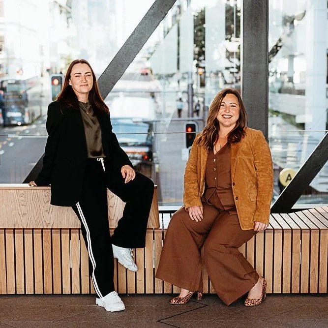 Two women sitting on a wooden bench in a modern indoor space with large glass windows and an urban cityscape background.