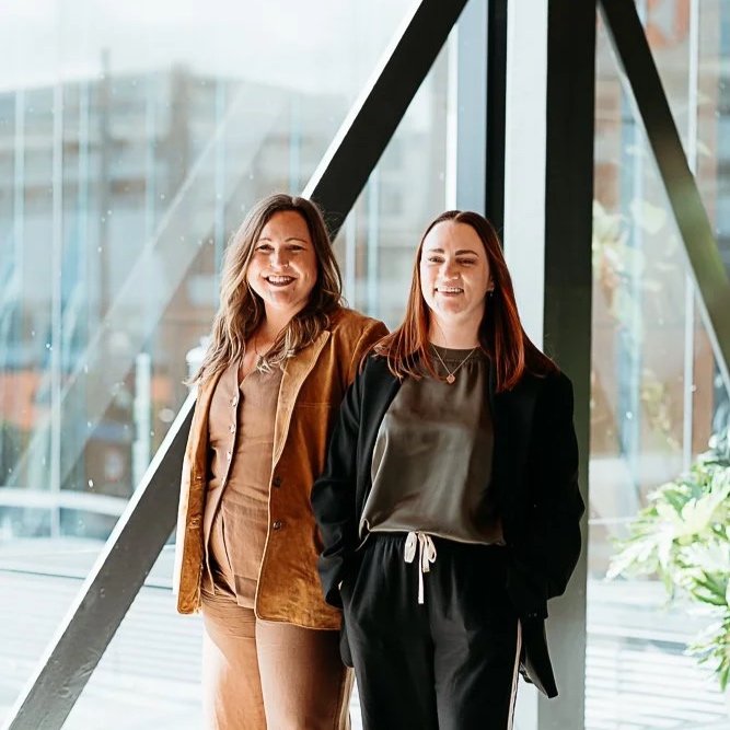 Two women standing inside a modern building with large glass windows, smiling and looking to the side.