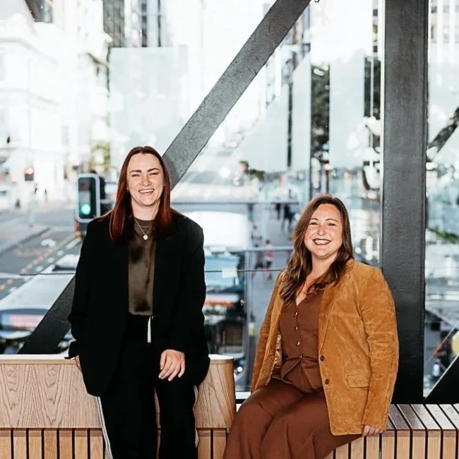 Two women smiling and posing in a modern urban setting with large glass windows and city streets in the background.