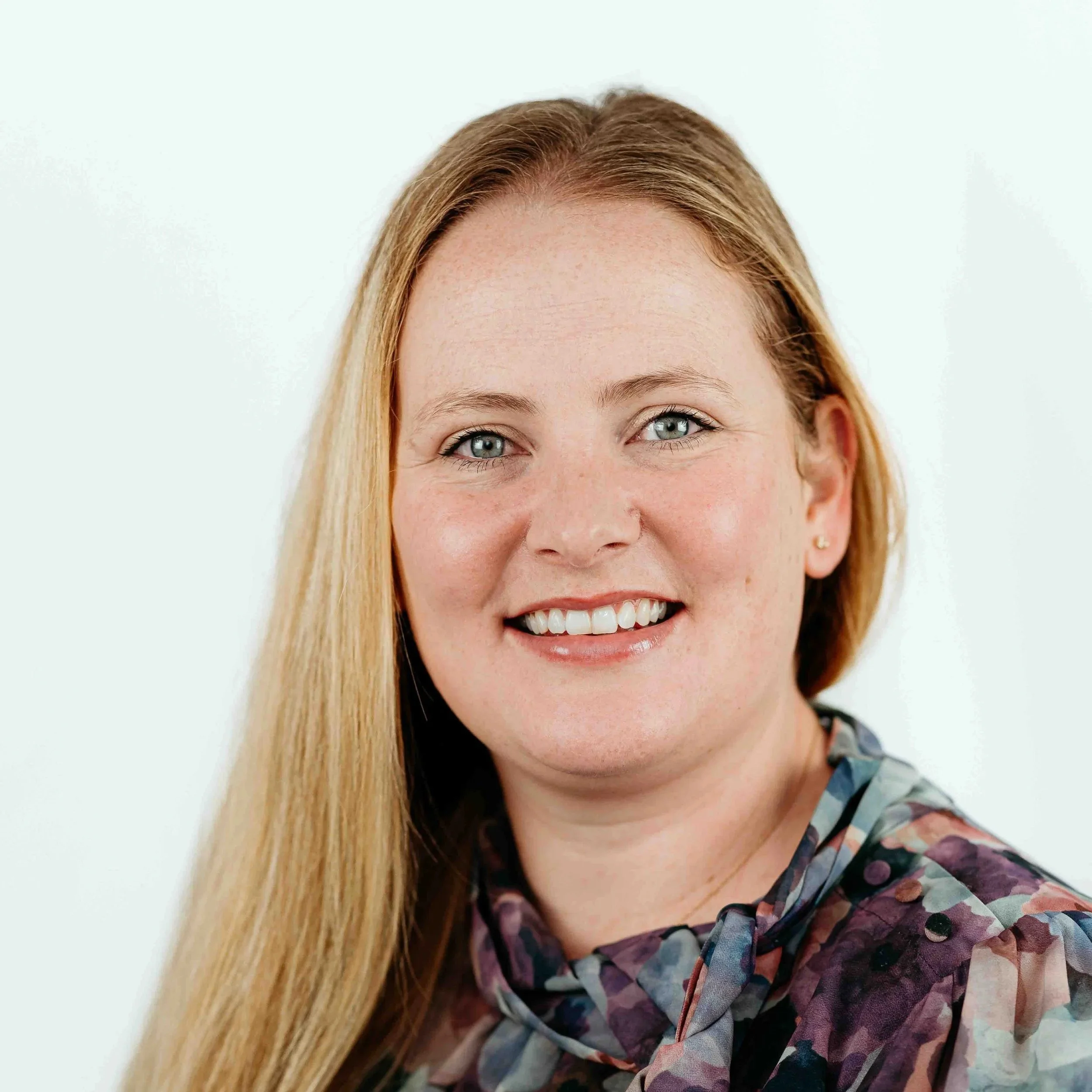 Close-up portrait of a smiling woman with long blonde hair, blue eyes, and light skin, wearing a multicolored patterned blouse with a bow at the collar.
