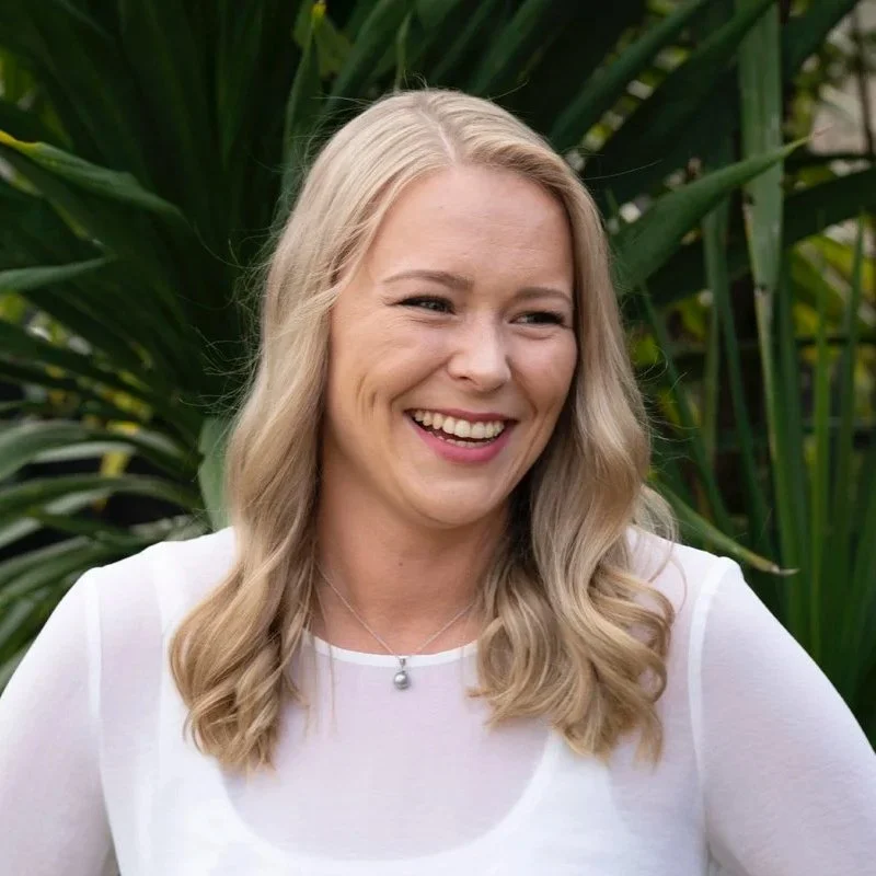 Smiling woman with long blonde hair wearing a white top, standing in front of green leafy plants.