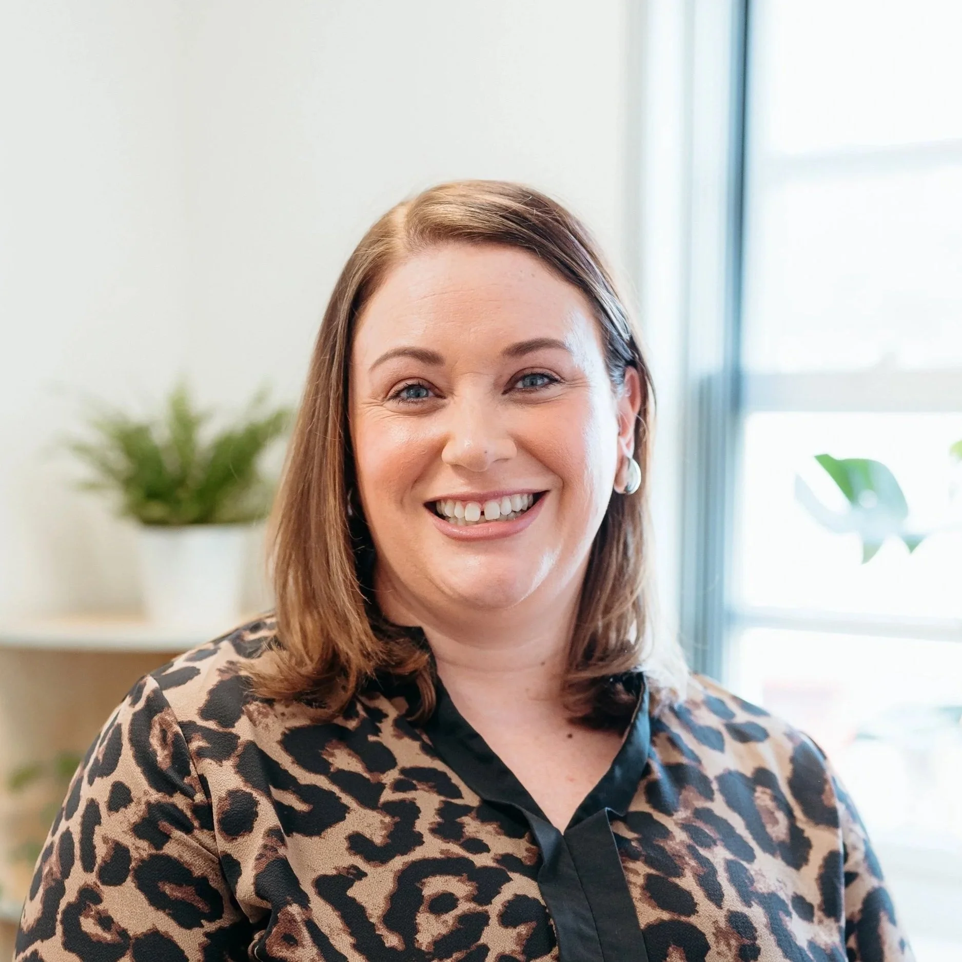 A woman with shoulder-length brown hair, wearing a leopard-print blouse, smiling in a well-lit indoor setting with plants in the background and a window with sunlight.