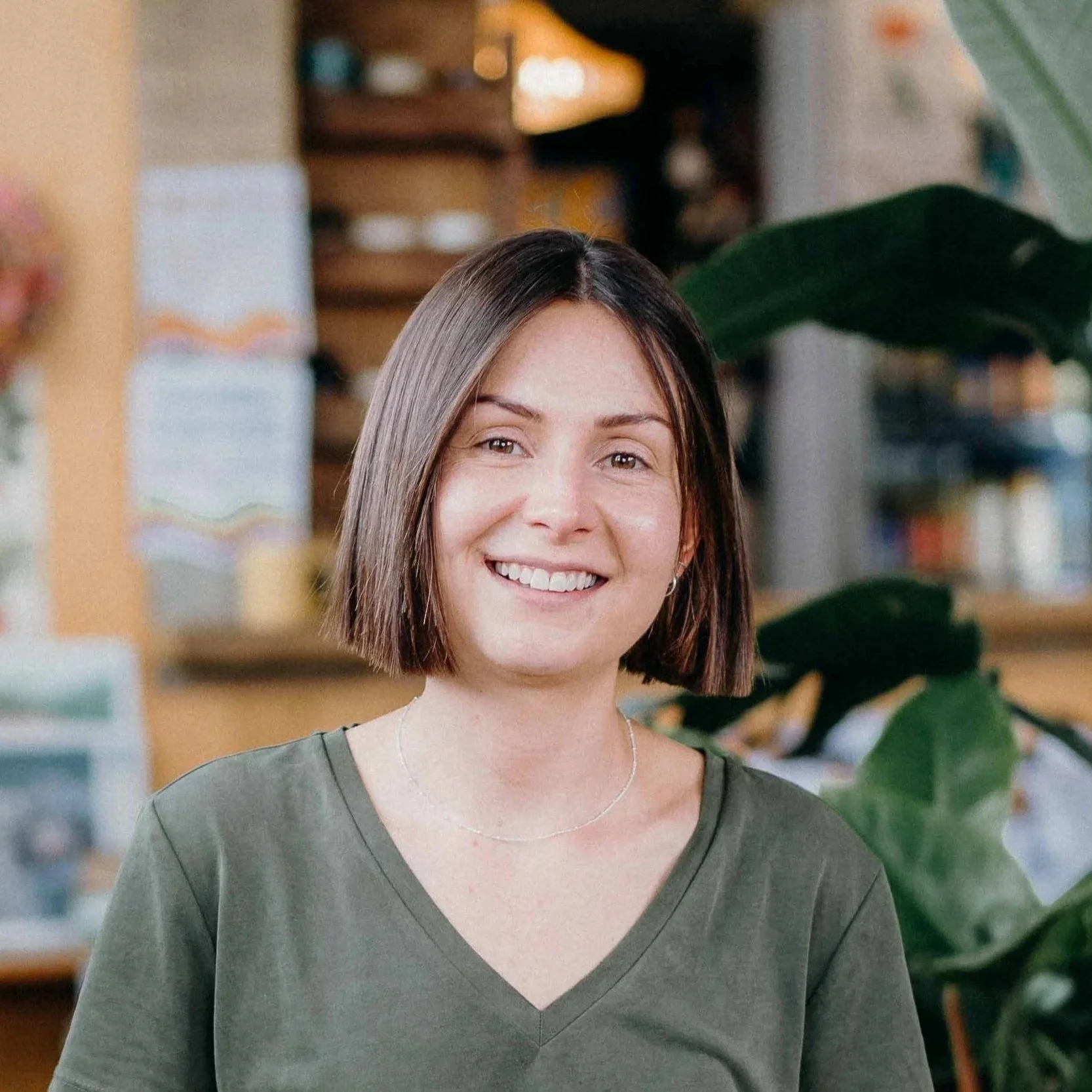 A woman with short brown hair wearing a green V-neck shirt smiling indoors with shelves and large green plant in the background.