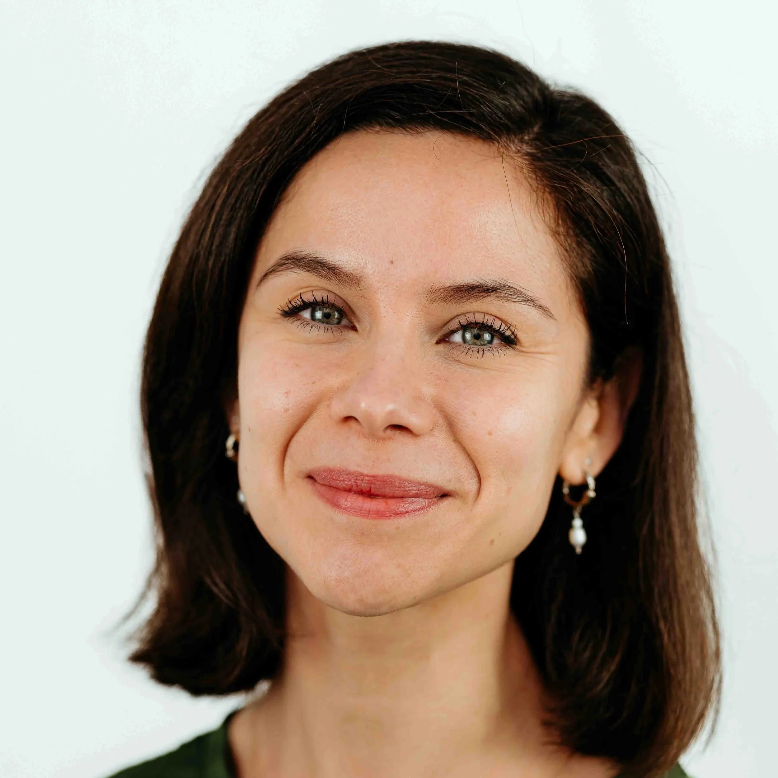 Close-up of a woman with brown hair, blue eyes, wearing pearl earrings, smiling against a white background.