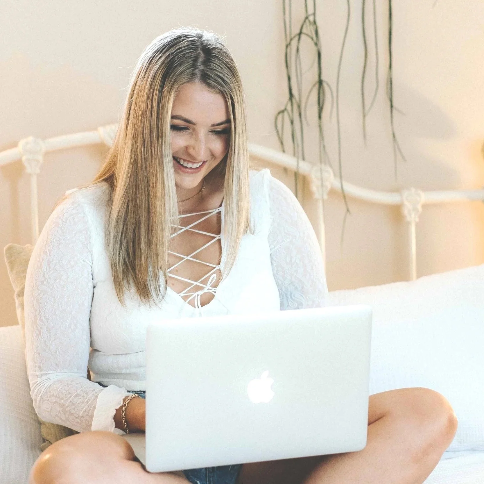 A young woman with blonde hair, wearing a white lace long sleeve top with criss-cross details on the front, sitting on a white couch while using a silver MacBook laptop, smiling at the screen in a brightly lit room with minimal decor and a potted plant in the background.