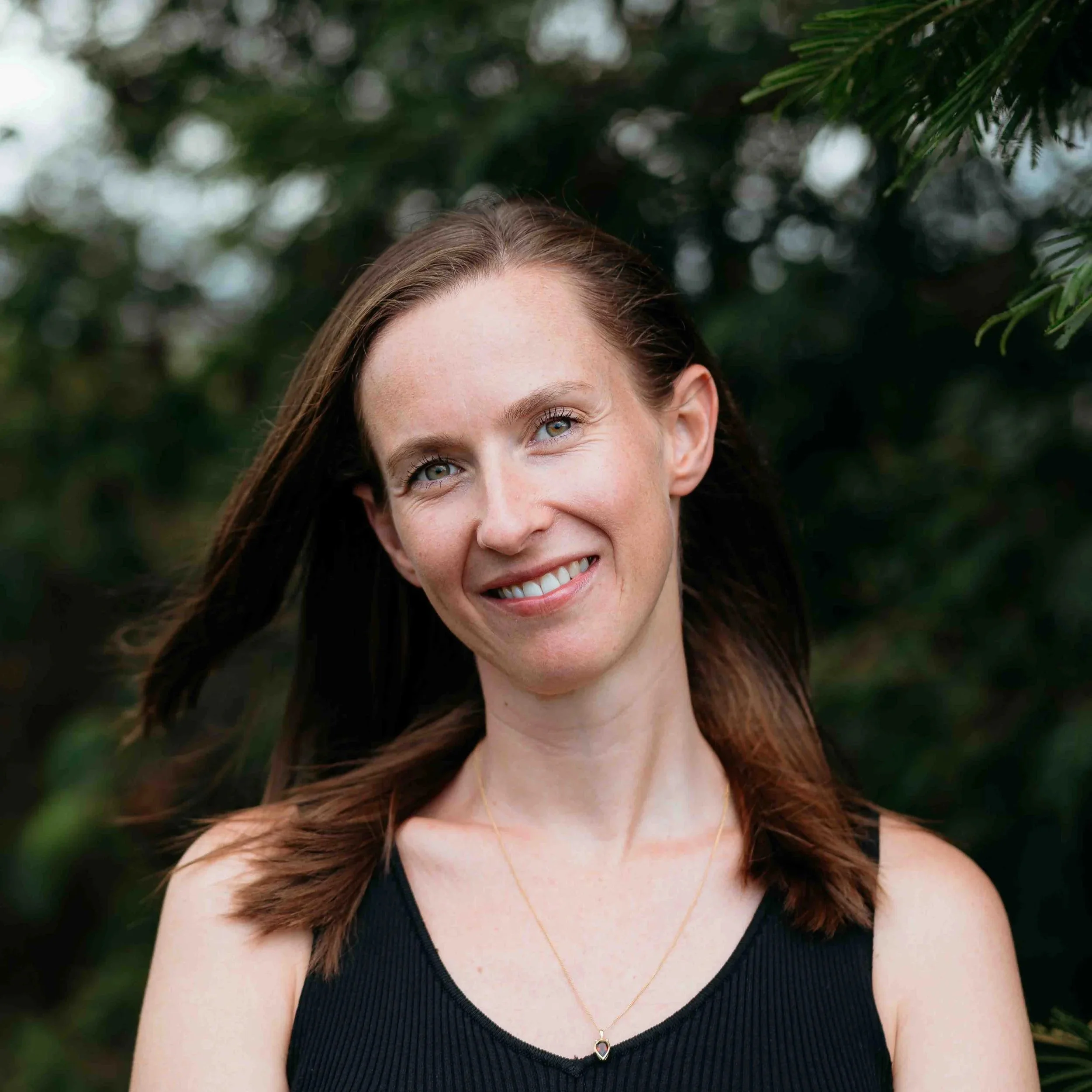 Woman in black sleeveless top smiling outdoors with trees in the background.